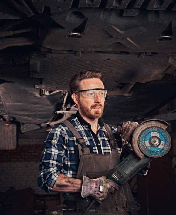 Two bearded auto mechanic working in repair garage. Auto mechanic in a uniform and safety glasses holds an angle grinder while standing under lifting car in a repair garage.