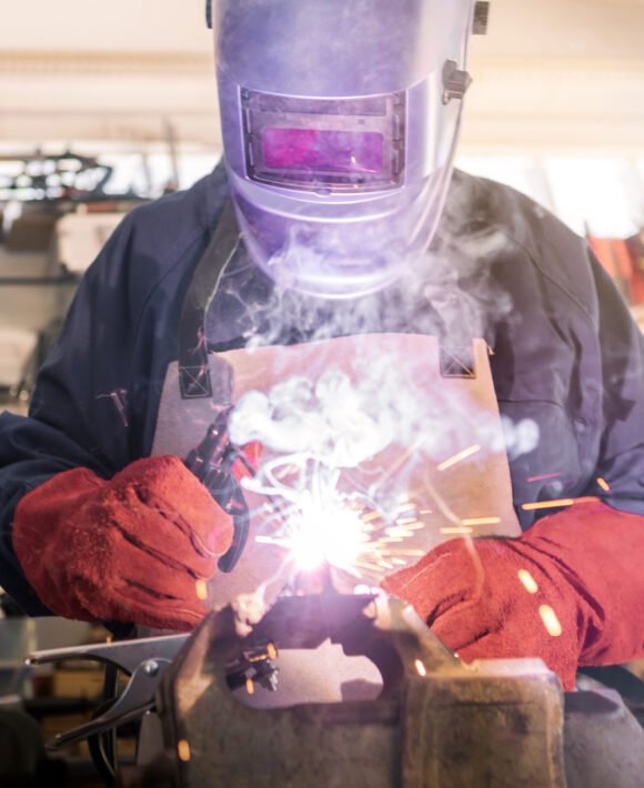 Worker in special clothes and helmet works as welding machine in his workshop Worker in special clothes and helmet works as welding machine in his workshop, bright sparks from metal heating and fly in different directions. The man works with an electric tool